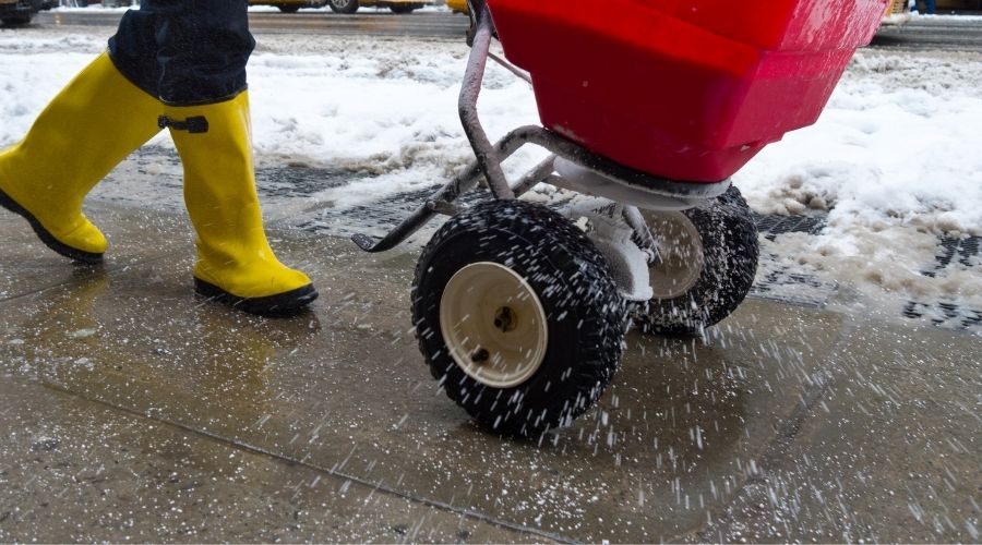 A person in yellow boots applies ice melt to a sidewalk.